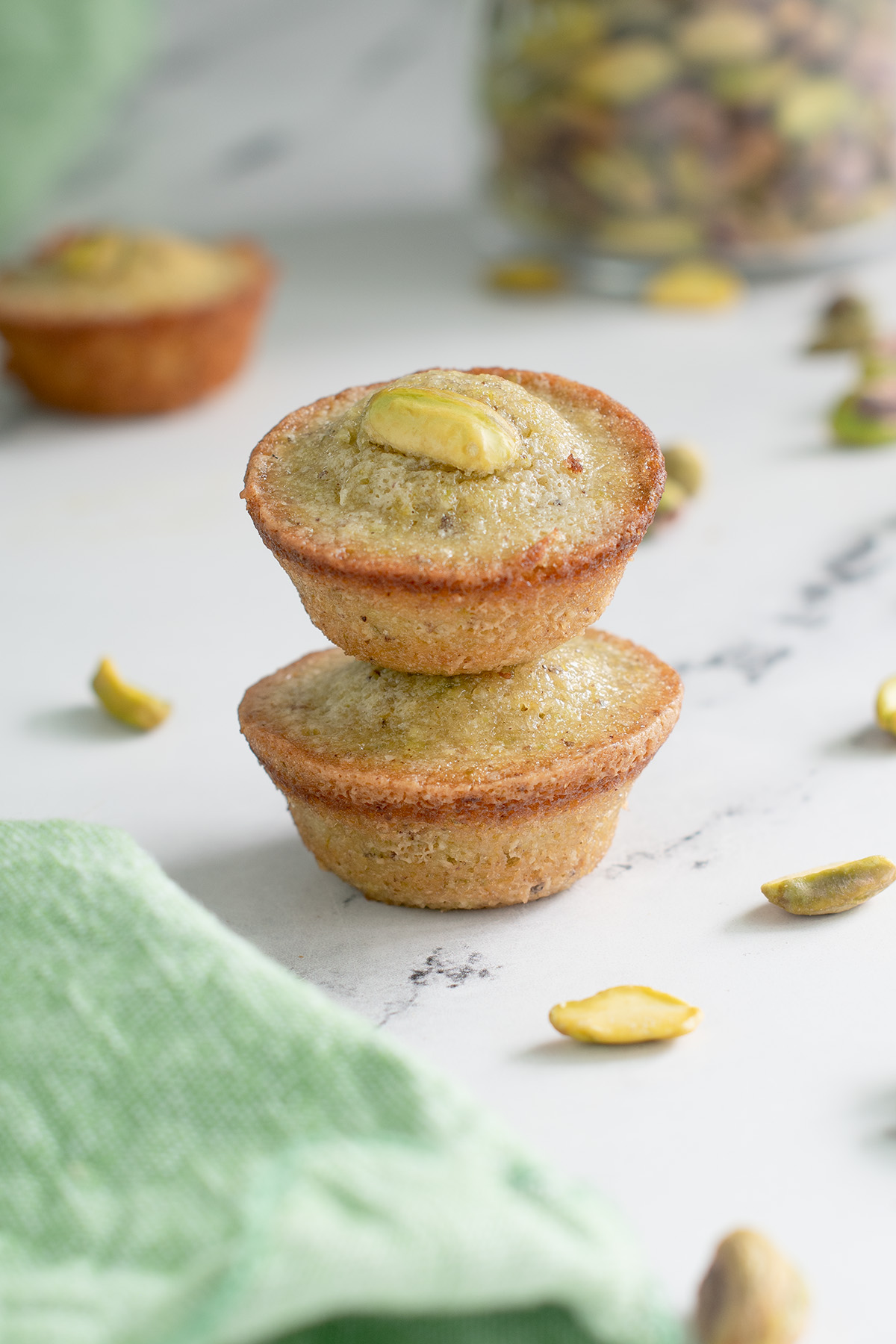 two pistachio financiers stacked on a white surface with a green cloth.