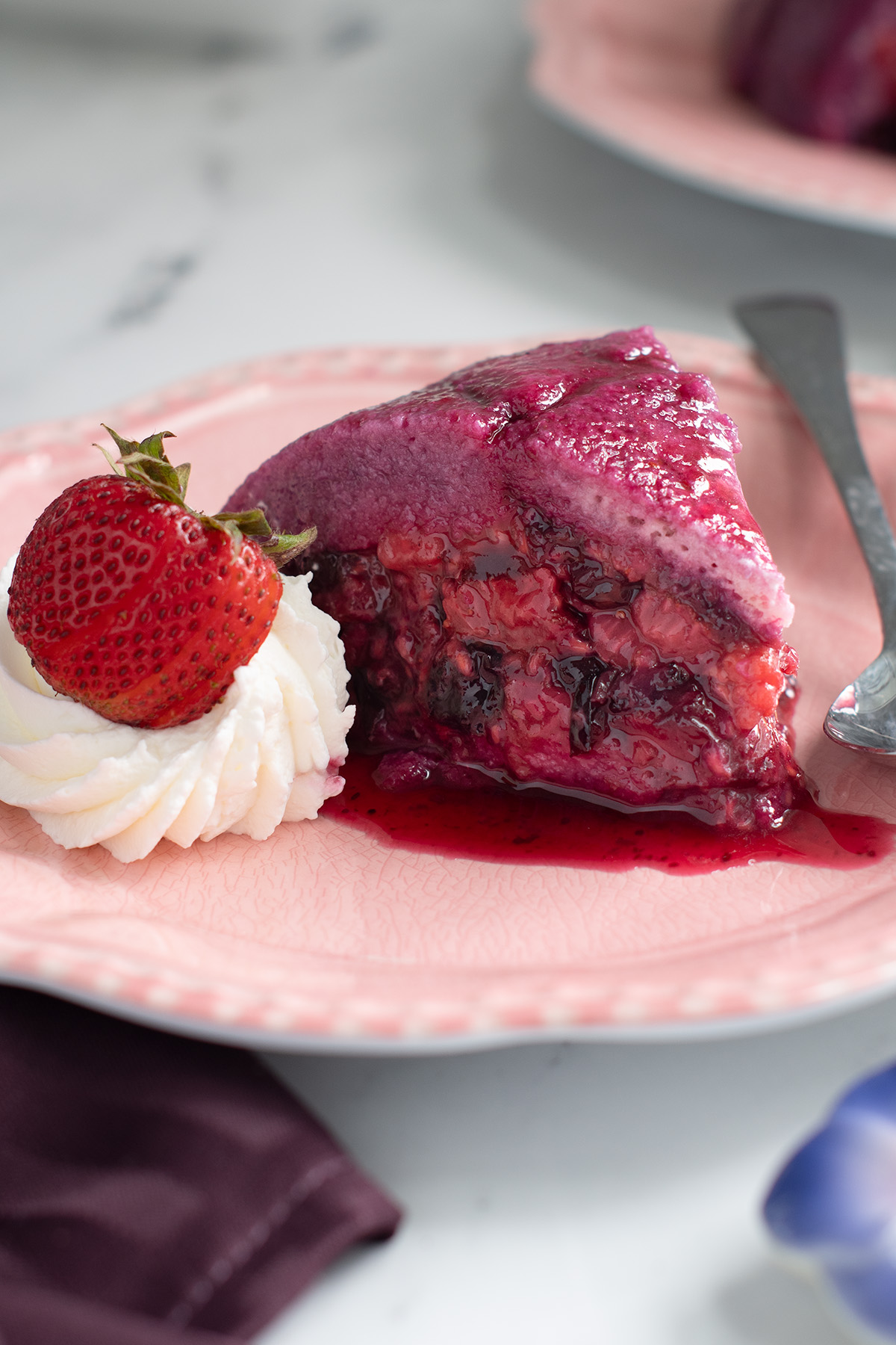 a slice of summer berry pudding on a pink plate with whipped cream and a spoon.