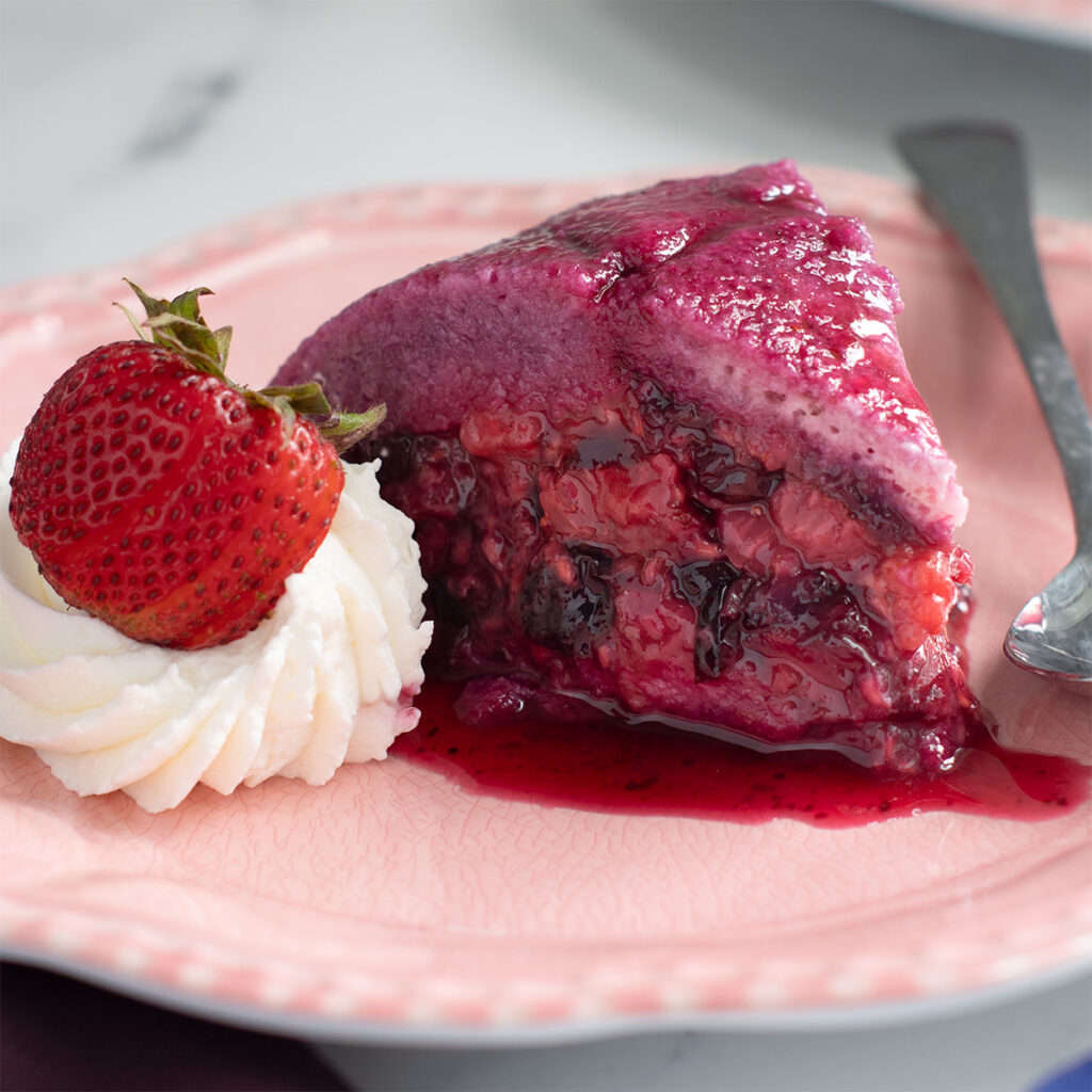 a slice of summer berry pudding on a pink plate with whipped cream and a spoon.
