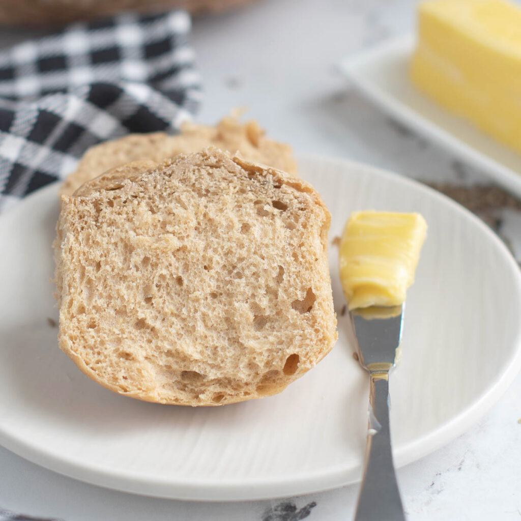 a split sourdough rye dinner roll on a white plate with a butter knife.
