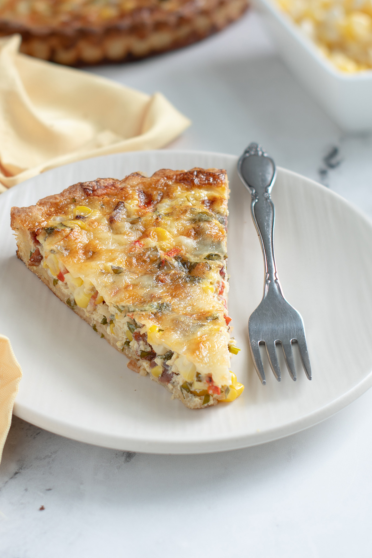a slice of corn quiche on a white plate with a fork and yellow napkin.