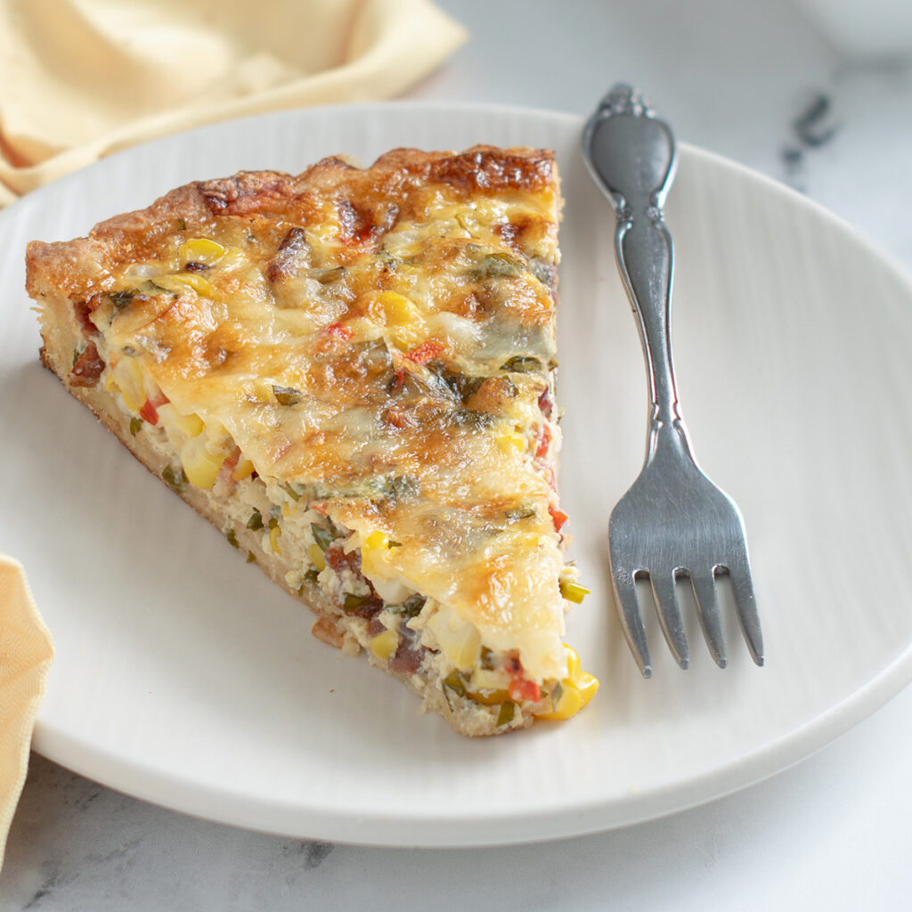 a slice of corn quiche on a white plate with a fork and yellow napkin.