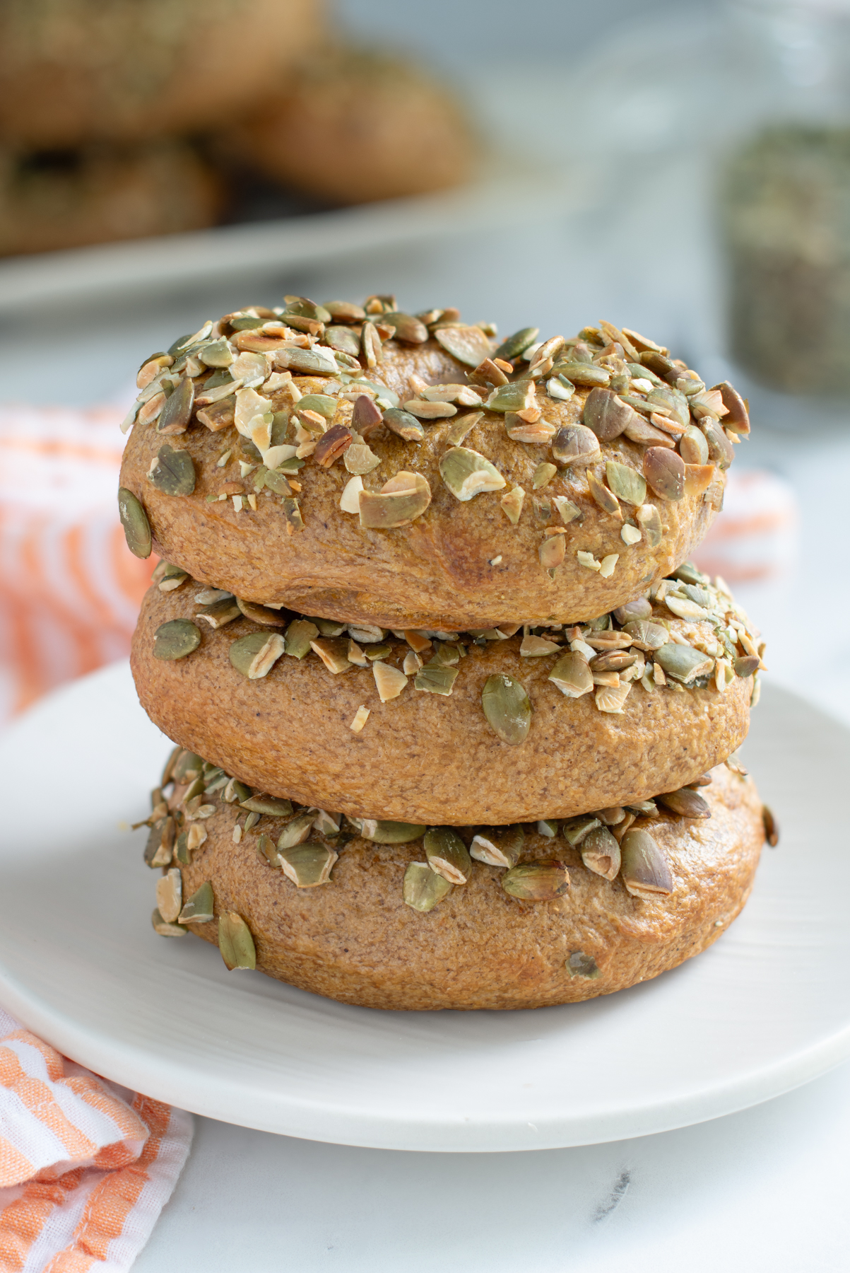 three sourdough pumpkin bagels with seeds stacked on a white plate.