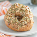 a sourdough pumpkin bagel on a white plate with seeds on top.