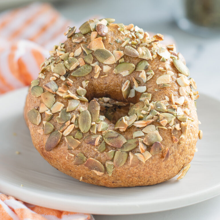 a sourdough pumpkin bagel on a white plate with seeds on top.